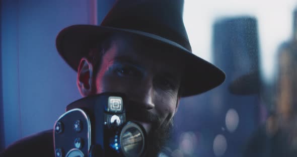 Man wearing hat looks around holding vintage camera in red blue lights alt