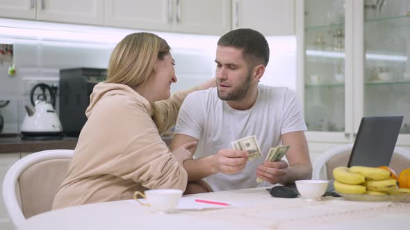 Portrait of Young Caucasian Husband Giving Money to Smiling Wife Sitting in Kitchen at Home alt