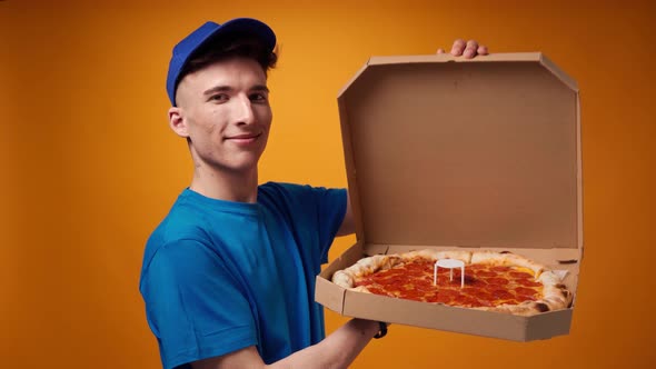 Young Deliveryman in Blue Uniform Opens a Box of Fresh Pizza Against Yellow Background