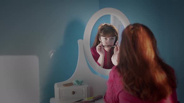 Woman Sitting in Front of a Mirror at Home Puts on Glasses and Smiles alt