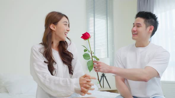 Asian young man surprise and give rose flower to girlfriend in bedroom. alt