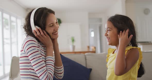 Happy hispanic mother wearing headphones listening to music and daughter sitting on sofa alt