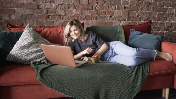 Blonde Lies on Red Sofa and Laughs Looking at Laptop Display
