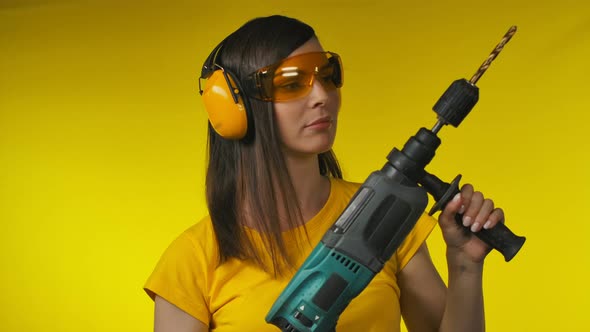 Young Brunette Woman in Protective Headphones and Glasses is Holding a Drill alt