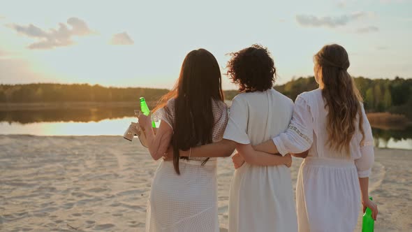 A Group of Female Friends Walks in an Embrace on a Lake Beach at Sunset