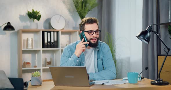 Man in Glasses Talking on Smartphone while Working on Laptop in Beautifully Decorated Room alt