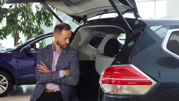 A Young Seller in a Suit Sits at the Open Trunk of the Car alt