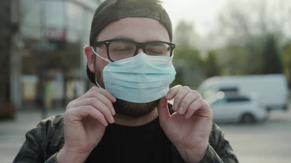 Young Man Wears Protective Medical Mask Corrects It and Looks at Camera Outdoors alt