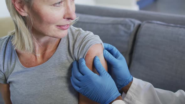 Happy senior diverse woman and doctor in living room sitting on sofa, vaccinating alt