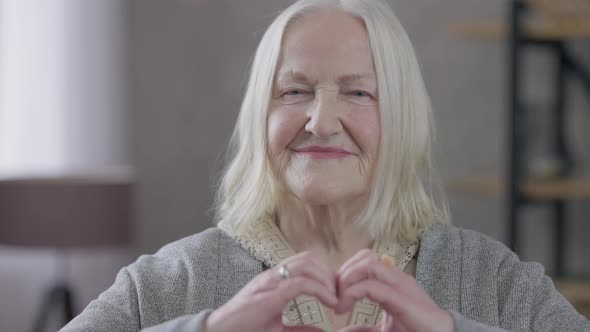 Old Greyhaired Caucasian Woman Showing Heart Shape with Hands Looking at Camera Smiling alt