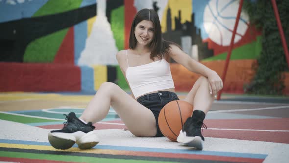 Smiling Confident Female Basketball Player Sitting on Outdoor Court with Orange Ball and Looking at alt