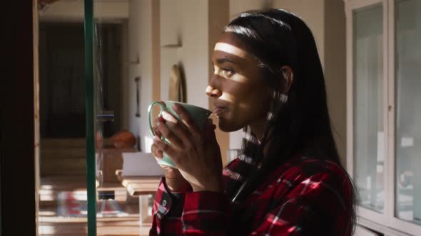 Mixed race woman standing by window at home having coffee alt