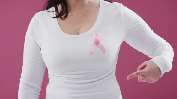 Mid section of a woman pointing to the pink ribbon on her chest against pink background alt