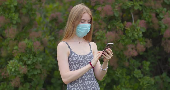 A young girl in a protective medical mask looks at the phone and sends messages