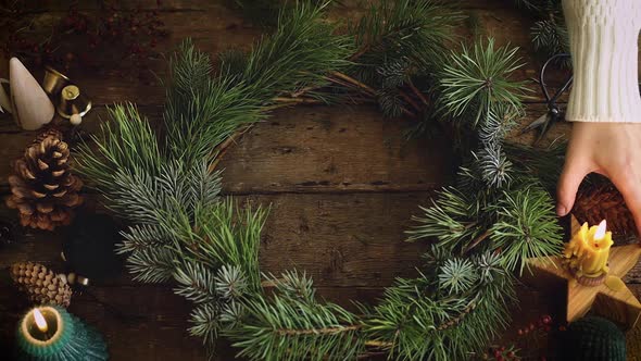 Hand putting burning candle in christmas wreath on rustic wooden table, top view alt