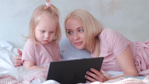 Young Attractive Blond Woman Teaches Her Little Charming Daughter in Pink Dresses Using a Tablet