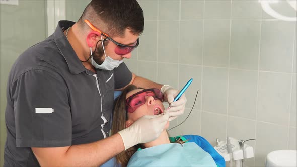 Doctor Orthodontist with Glasses Whitens the Teeth of a Young Woman with a Laser alt