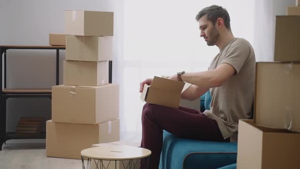 A Young Man Opens a Cardboard Box and Takes Out Things Sitting on the Sofa in a New Apartment After alt