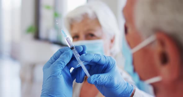 Male caucasian doctor holding syringe while senior caucasian woman wearing face mask sitting with sl alt