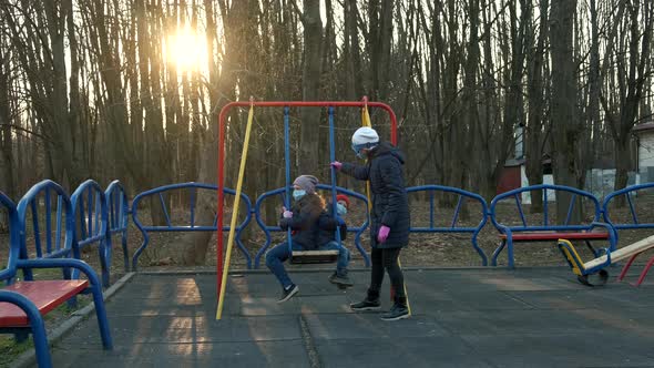 Mother and children in medical masks swing on a swing in the Playground .