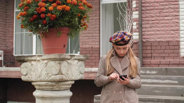 Young Girl in Coat and Beret Using Smartphone on Stairs Brick House in Garden. Beautiful Girl alt