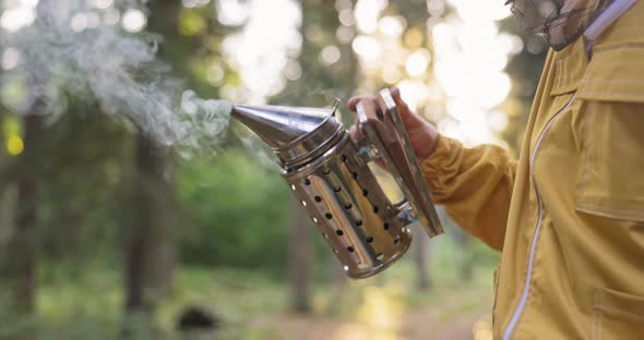 A Beekeeper Dressed in a Protective Suit with a Mosquito Net Over His Head Sprays White Smoke Over alt
