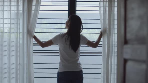 Attractive young Asian woman opening curtains in the room at home.