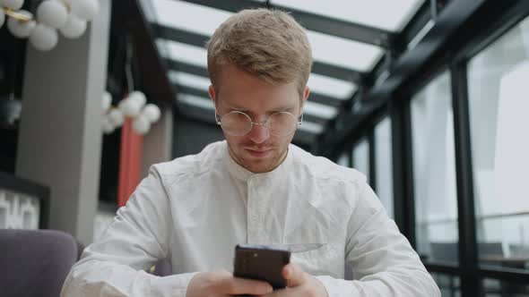 Handsome Caucasian Man with Eyeglasses is Using Smartphone for Chatting and Sending Email Sitting alt
