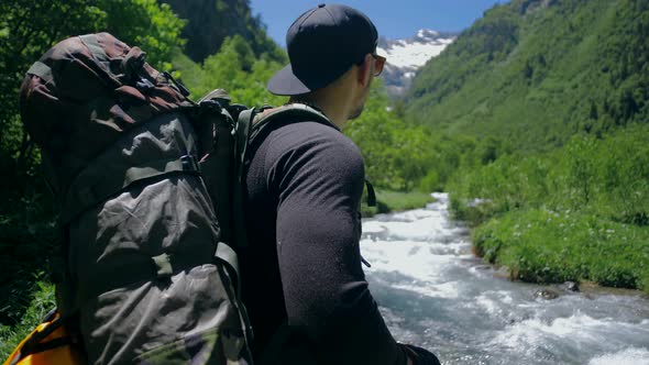 Traveler with a large backpack is standing by the mountain river alt