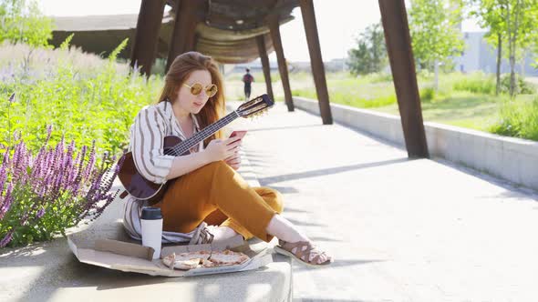 Woman with Ukulele Surfs Internet with Smartphone in Park