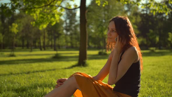 Happy Attractive Sexy Lady Sitting on Green Grass and Have Conversation Via Digital Phone. alt
