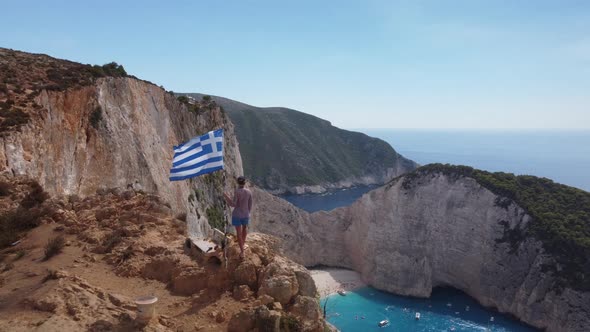 Caucasian woman with greek flag on Zakynthos beach cliff. Aerial drone view alt