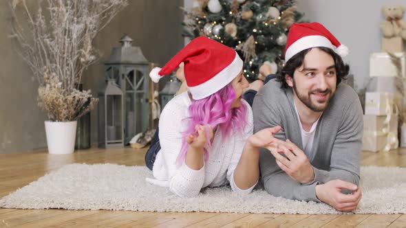 Happy Young Couple in Santa's Red Hats Is Lying Together Near Christmas Tree.
