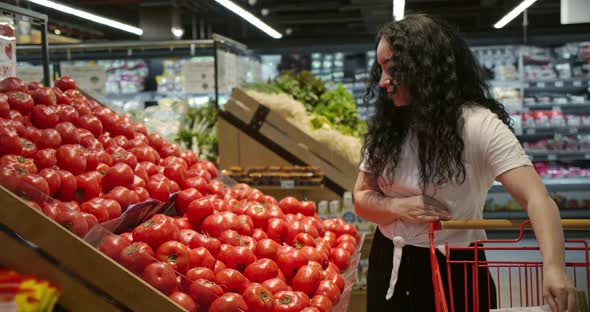 Young Woman in the Grocery Section of a Supermarket Picks Up a Bag and Puts Tomatoes in a Basket to alt