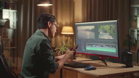 Asian Male Video Editor Works And Using Mobile Phone With His Personal Computer alt