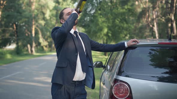 Drunk Young Man Drinking Wine From Bottle Leaning on Car Standing at Suburban Road alt