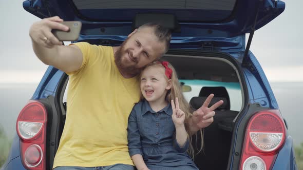 Cheerful Young Father and Cute Daughter Taking Selfie on Smartphone Sitting in Car Trunk Grimacing alt