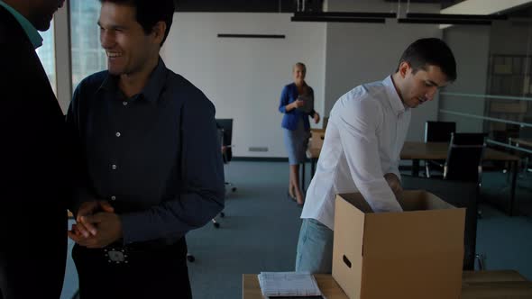 Young Employee Unpack Box with Docs and Equipment. His Colleagues Walking Near in Modern Office alt