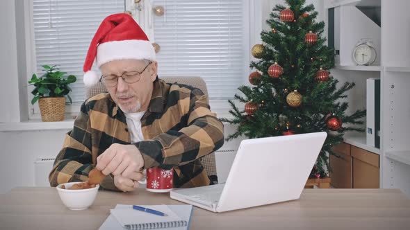 A Middleaged Man in a Santa Claus Hat Drinks a Hot Drink From a Cup and Eats Cookies While Sitting