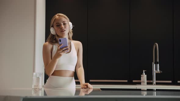 A Young Blonde in Headphones Dancing in the Kitchen Happily Listens to Music alt