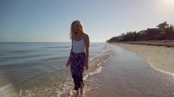Pretty, carefree woman in sarong and tank top splashing in the water walking on the beach in slow mo alt
