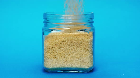 Closeup of Falling Down Wheat Into Glass Jar on Blue Background alt