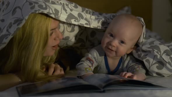 woman plays with her baby son under the covers in the evening in the bedroom