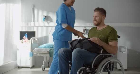 Afro Female Nurse Talking to Discharged Male Patient in Wheelchair and Smiling alt