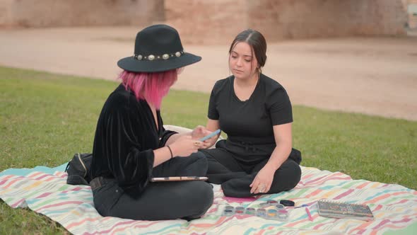 Two Friends in Black Clothes Sit on Ground and Interact While One Texts on Cellphone alt
