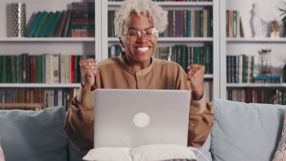 Young African American Woman with Laptop on Lap Overjoyed By News Sits on Sofa alt