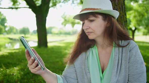 Brunette Woman in a Hat Sits in the Park Under a Tree and Uses the Phone