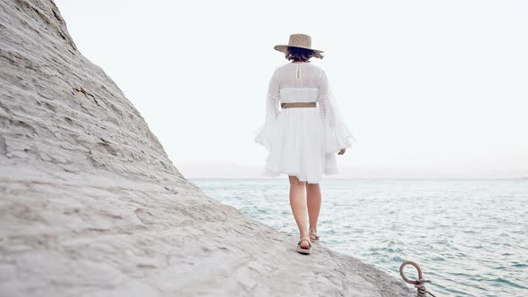 Stranger Woman in White Dress Walking on Clay Beach Near Mediterranean Sea alt