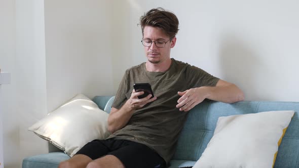 Young Modern Guy Communicates on the Phone While Sitting in the Room on the Couch alt