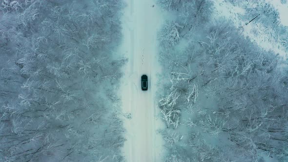 Aerial View of a Car Driving on a Snowy Road in a Winter Forest alt